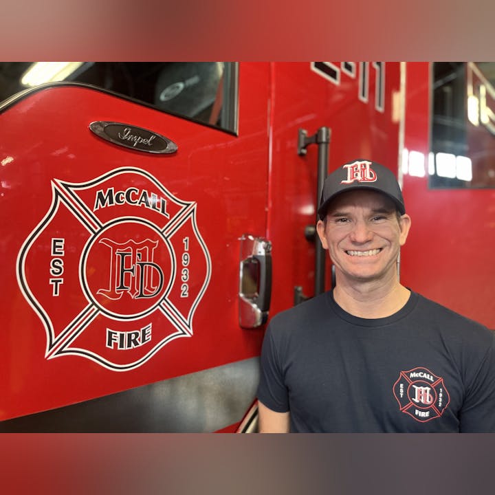 A smiling person wearing a cap and t-shirt with a fire department logo, standing in front of a red fire truck with similar insignia.