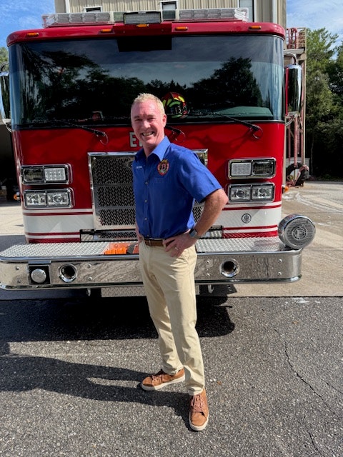 A person in a blue shirt stands in front of a red fire truck on a sunny day.