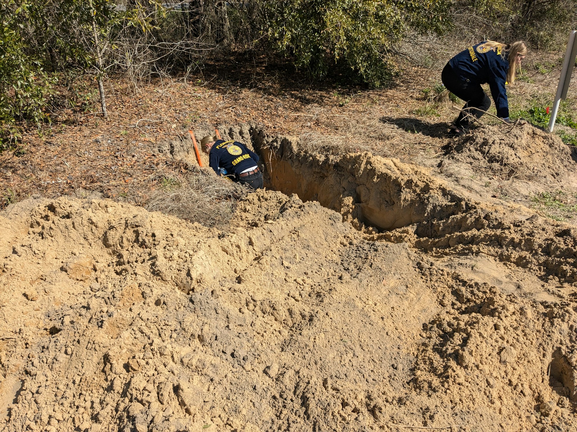 Two people digging in the soil, wearing matching uniforms, surrounded by dirt piles and trees.