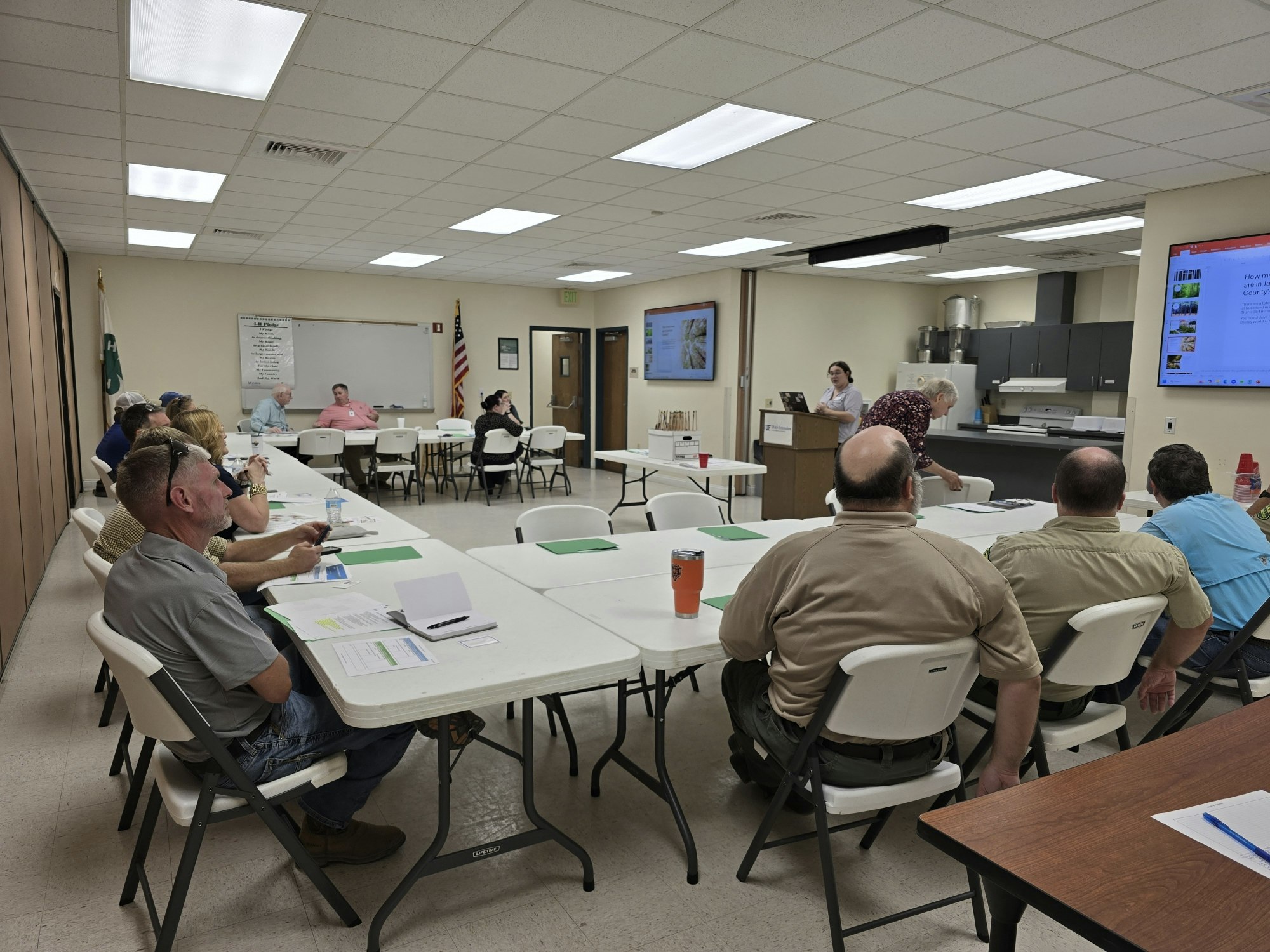 A group of people in a conference room attending a presentation, with notes and drinks on tables.