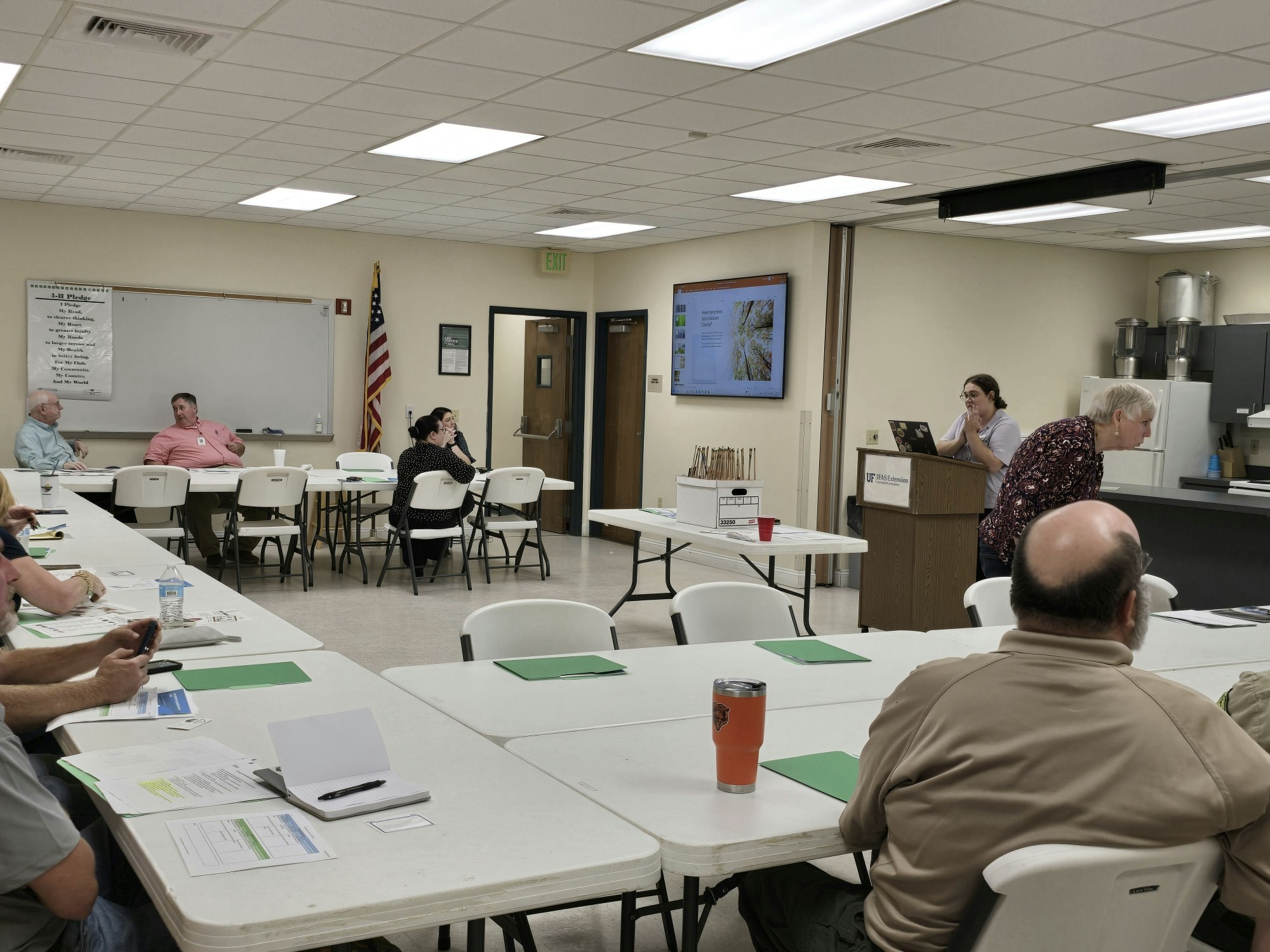 A classroom with people seated at tables, listening to a presentation. One person is at a podium, and slides are shown on a screen.