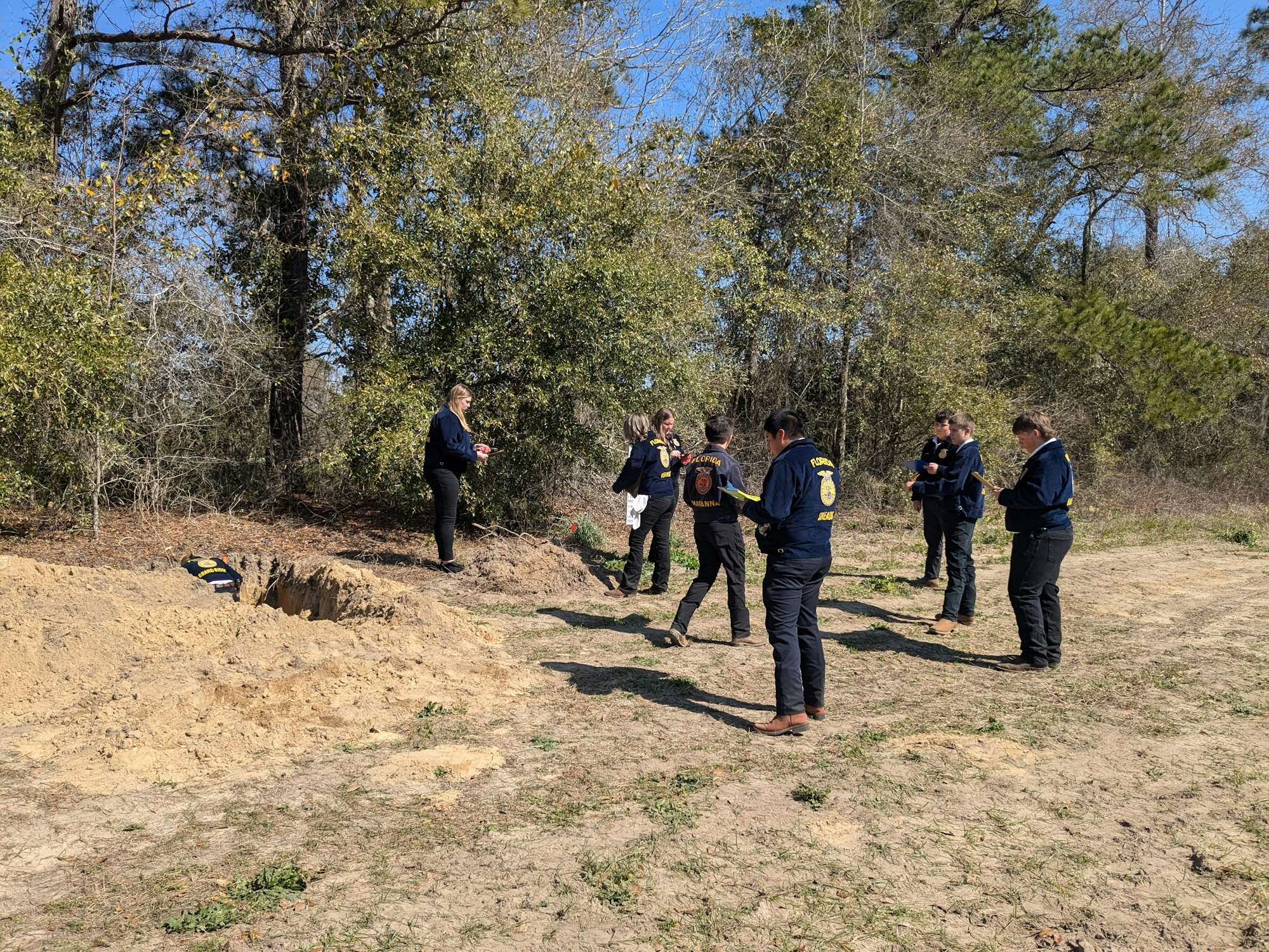A group of people in matching jackets are gathered around a hole in a wooded area.