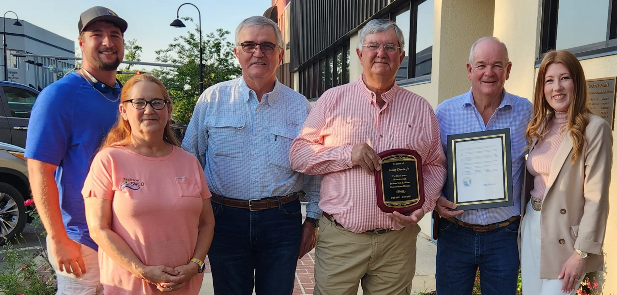 A group of six people gathered outside, with two men holding awards, celebrating a recognition event.