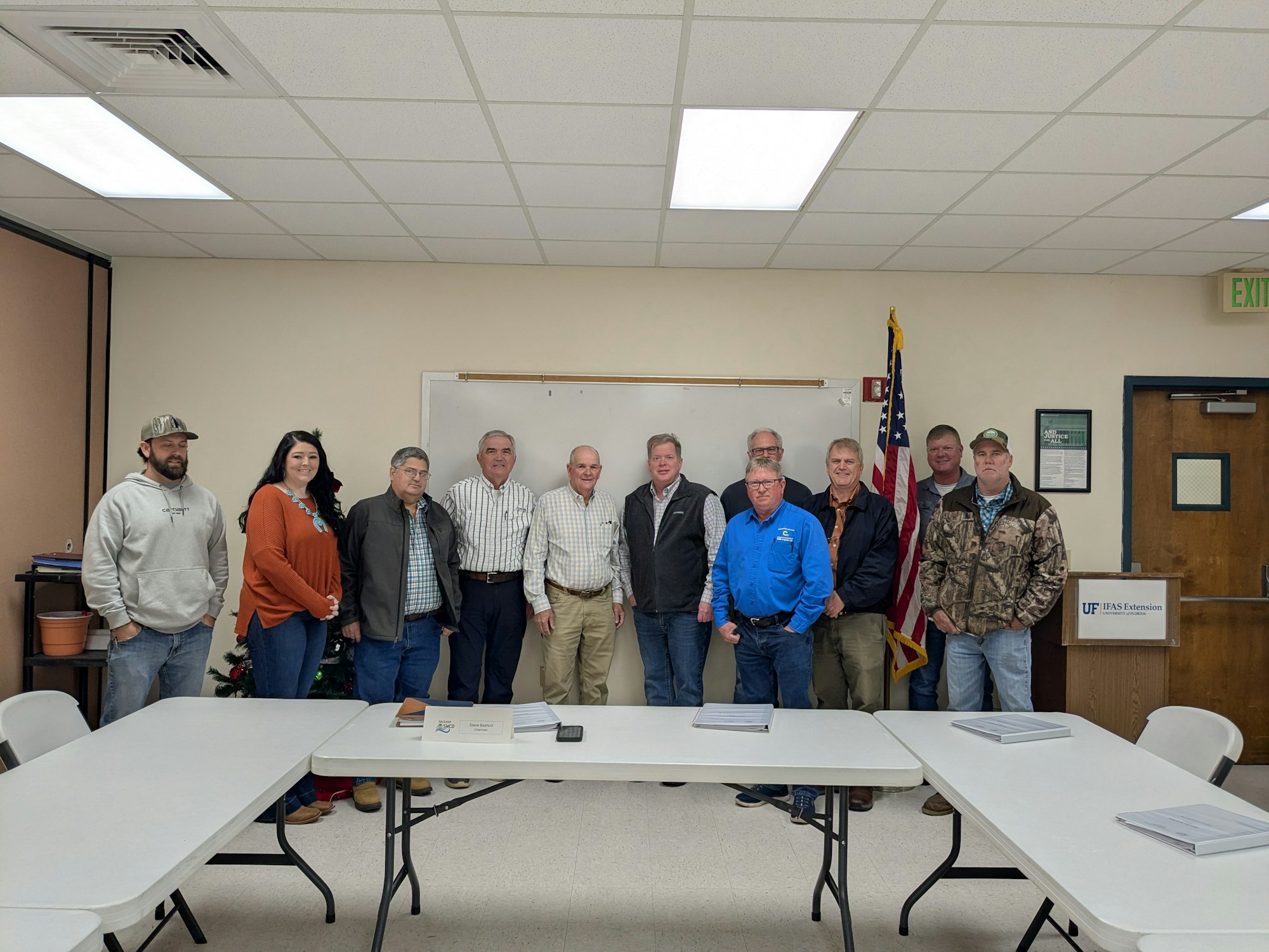 A group of people posing for a photo in a classroom setting, with a Christmas tree and an American flag visible.