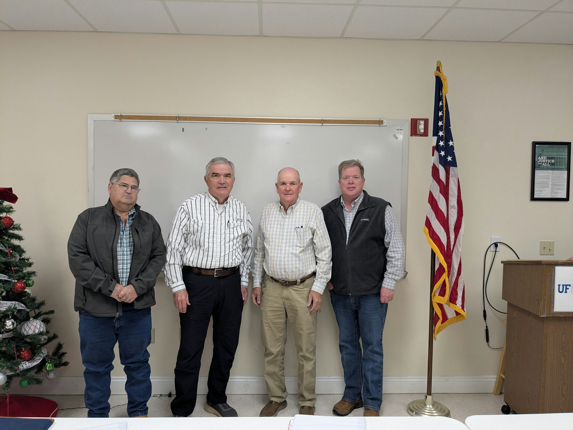 Four men posed in a room with a whiteboard and an American flag, next to a small Christmas tree.