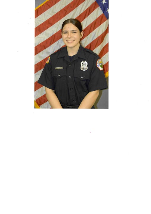 A person in a uniform stands in front of a U.S. flag, smiling.