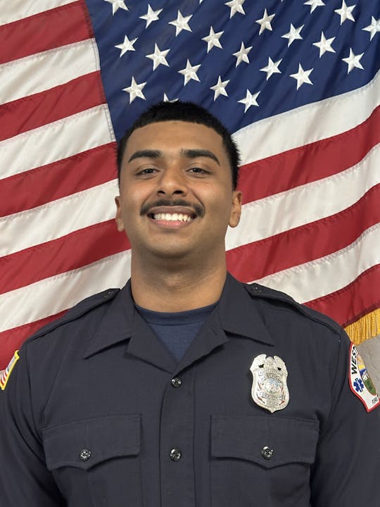 A person in a uniform poses in front of an American flag backdrop, smiling.