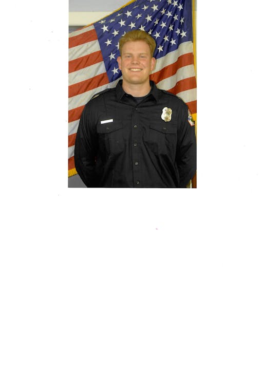 A person in a uniform stands in front of a U.S. flag, smiling.