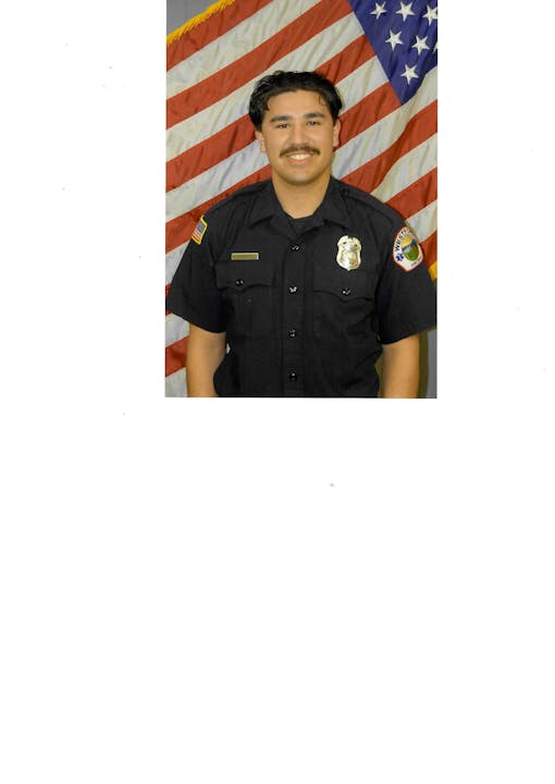 A person in a black uniform stands in front of an American flag, wearing a badge and nameplate.