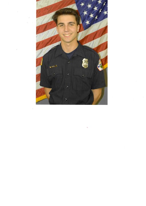 A person in a dark uniform stands smiling in front of an American flag backdrop.