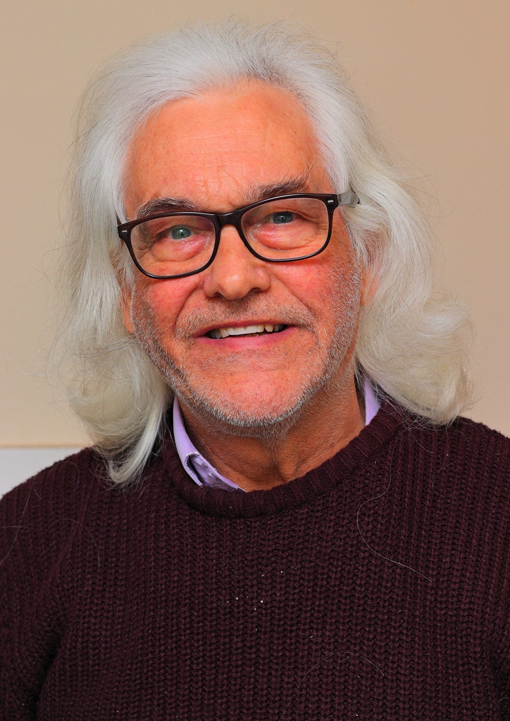 An older person with gray hair and glasses, wearing a dark sweater, smiling.