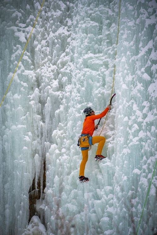 Person ice climbing on a frozen waterfall with climbing gear and orange clothing.