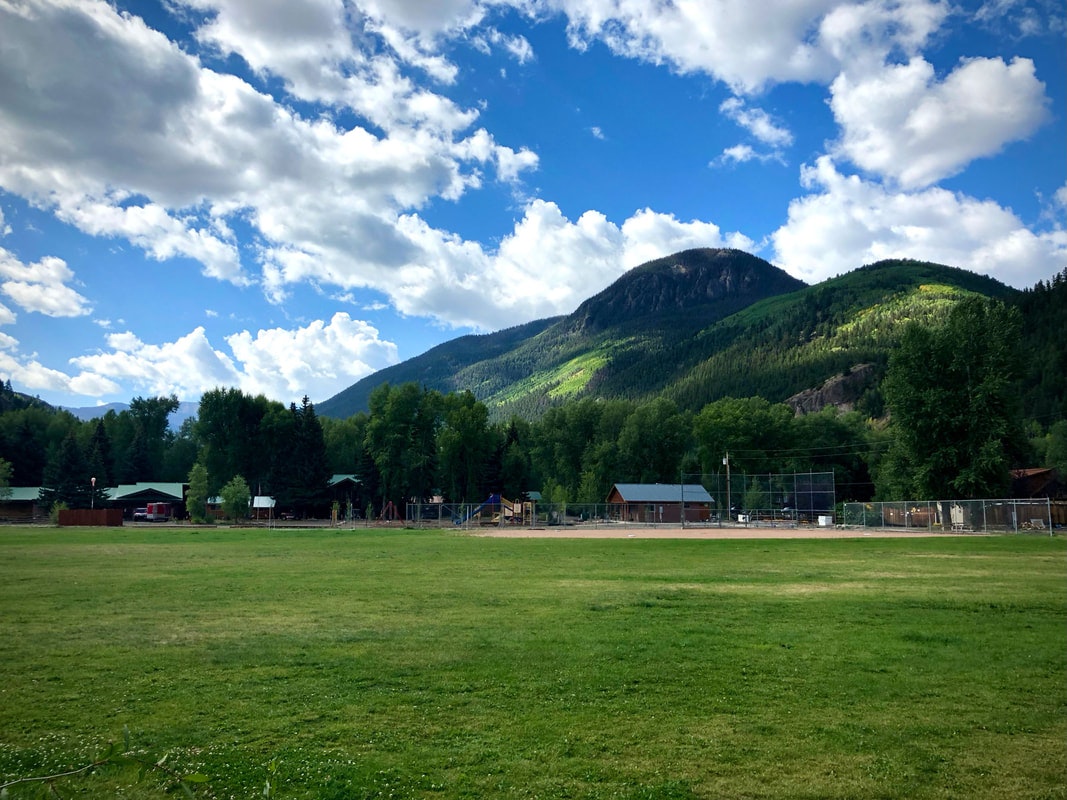 A grassy field with buildings, trees, and a mountain in the background under a partly cloudy sky.