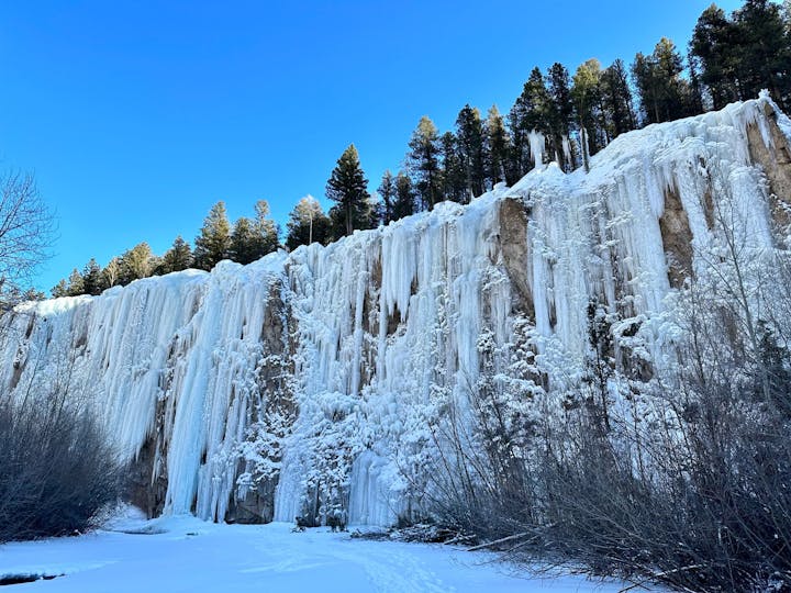 A frozen waterfall cascades over a rocky cliff surrounded by snow, with pine trees atop under a clear blue sky.