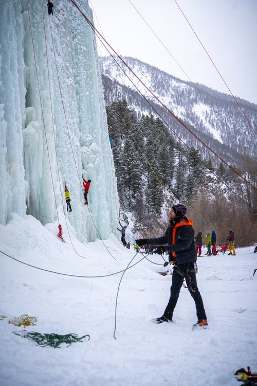 People ice climbing a frozen cliff with ropes and harnesses in a snowy mountain setting.