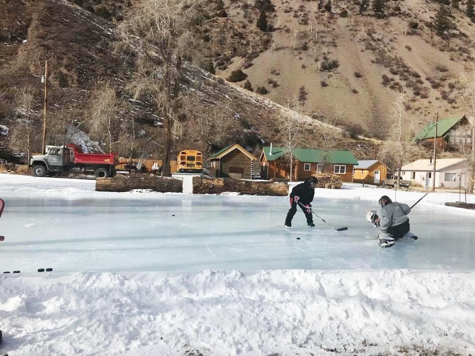 Two people play ice hockey on a snowy outdoor rink with a scenic mountain backdrop and cabins.