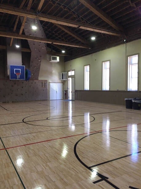 An indoor basketball court with a climbing wall, wooden floor, and high ceiling.