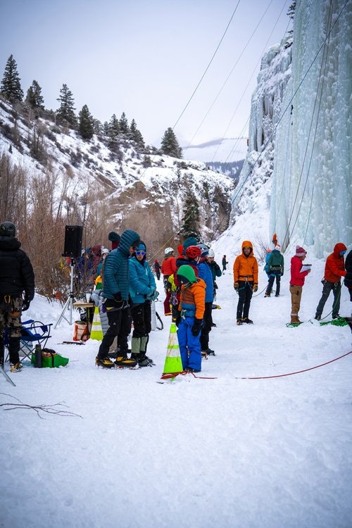 People in winter gear gathering near an ice-covered cliff with climbing gear in a snowy landscape.
