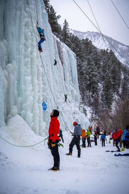 People ice climbing on a frozen waterfall, with others watching and belaying. Snowy landscape with trees and mountains in the background.