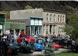 A crowd gathers around vintage cars in a historic town setting with old buildings.