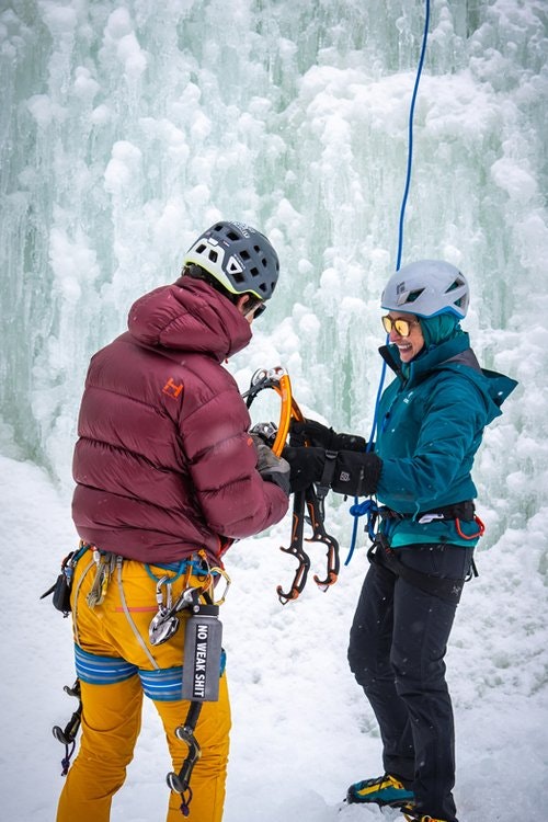 Two people in helmets and gear prepare for ice climbing on a snowy surface.