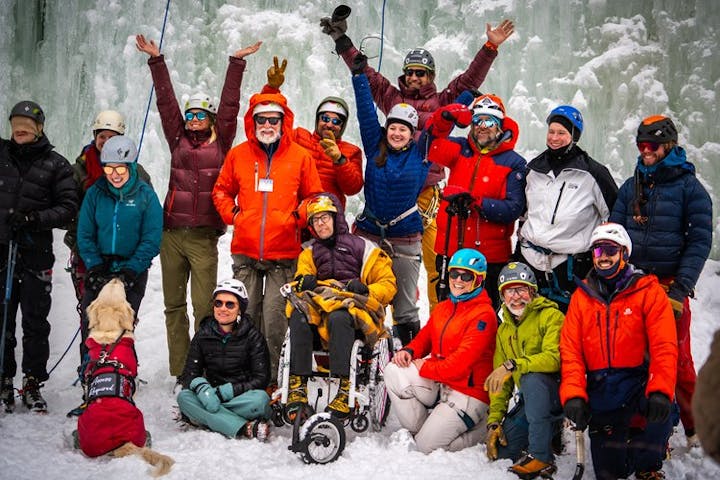 A group of people in winter gear, some in wheelchairs, smiling and raising hands, with an ice wall in the background. A service dog is present.
