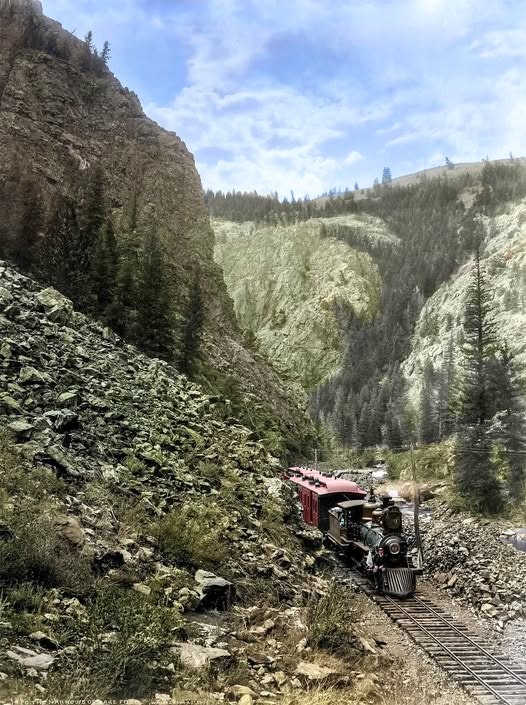 A vintage train navigates a rocky mountain landscape, surrounded by tall trees and steep cliffs under a cloudy sky.