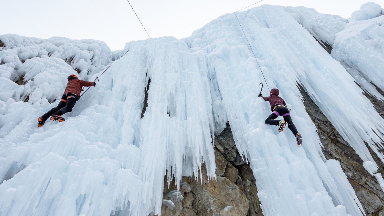 Two people are ice climbing a frozen waterfall with ice axes and crampons.