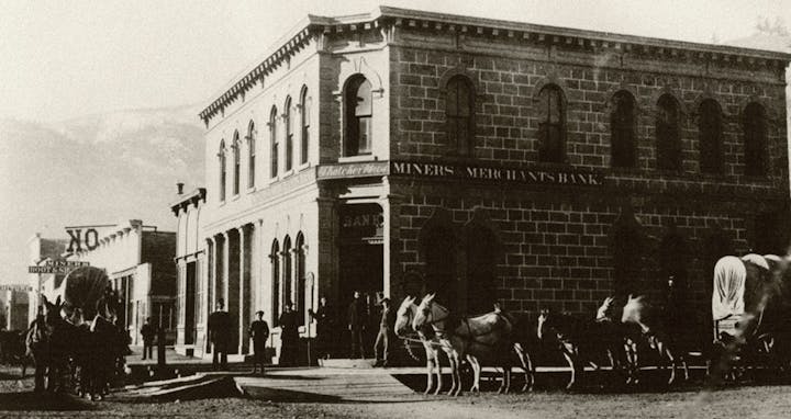 Historic scene with brick bank building, horses, carriages, and people on a street. Signs say "Miners Merchants Bank" and "Boot & Shoe."