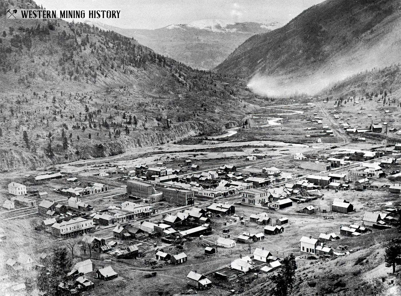 A historical black-and-white photo of a mining town nestled in a valley, surrounded by mountains and featuring numerous buildings.