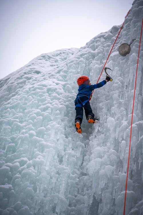 Person in blue gear ice climbing with an ice axe on a frozen wall, wearing a red helmet and crampons, attached to a red rope.