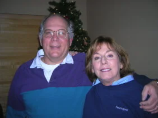 Two smiling people in layered clothing with a Christmas tree in the background.