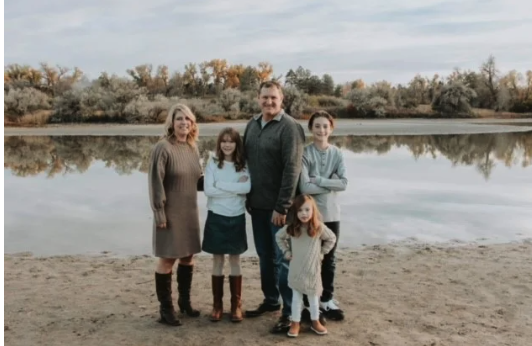 A family of five stands smiling by a tranquil lake with autumn trees reflected in the water.