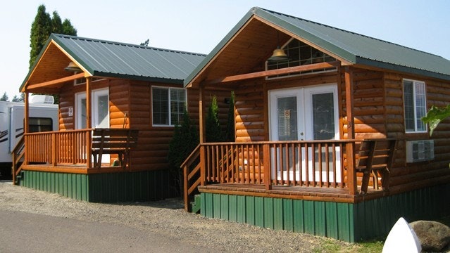 Two small wooden cabins with green roofs, each with a porch and railings, set on a paved path surrounded by greenery.
