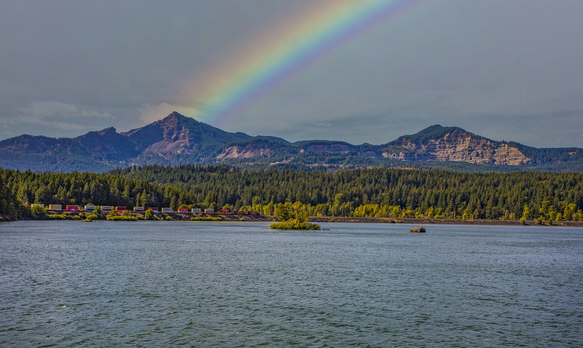 ©RHALLMAN; A vivid rainbow arching over a landscape with a river, forest, mountains, and a colorful train.