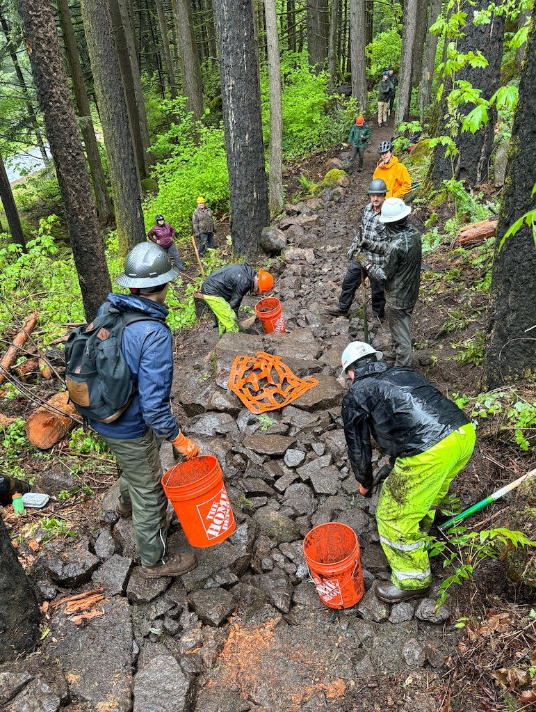 People in a forest wearing helmets, working on a rocky path with orange buckets.