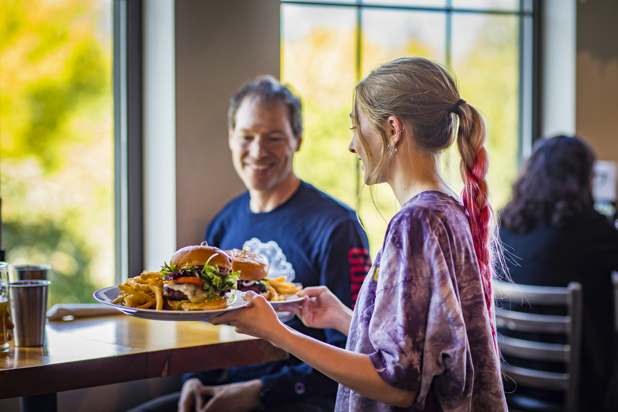 A woman serves burgers and onion rings to a man at a restaurant table, with a bright window in the background.