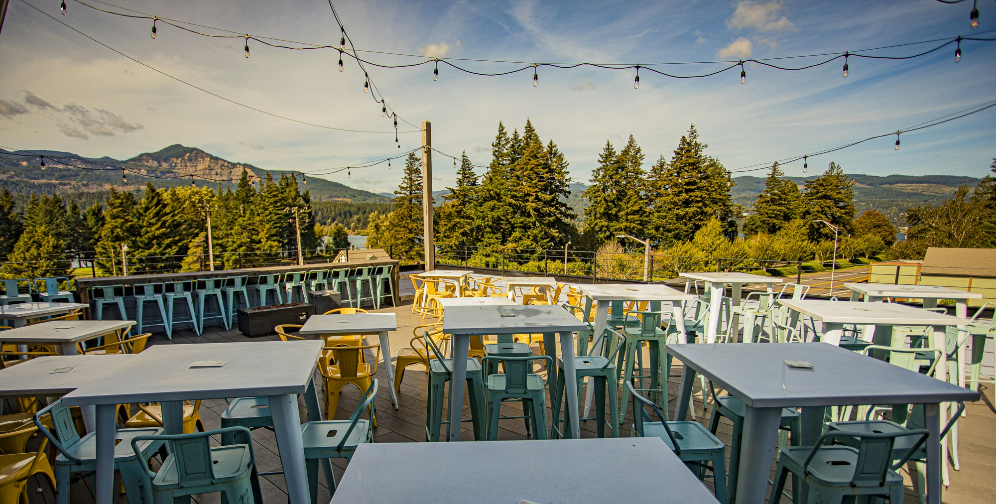 Outdoor seating with tables and chairs under string lights, mountains and trees in the background.
