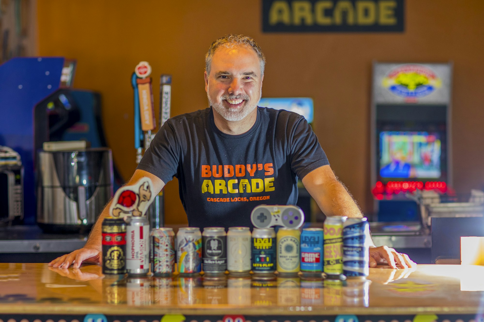 Smiling person at an arcade counter with various beer cans displayed in front.