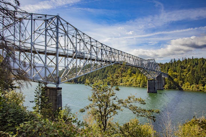 ©RHALLMAN; A steel truss bridge spanning a river, surrounded by trees under a cloudy sky.