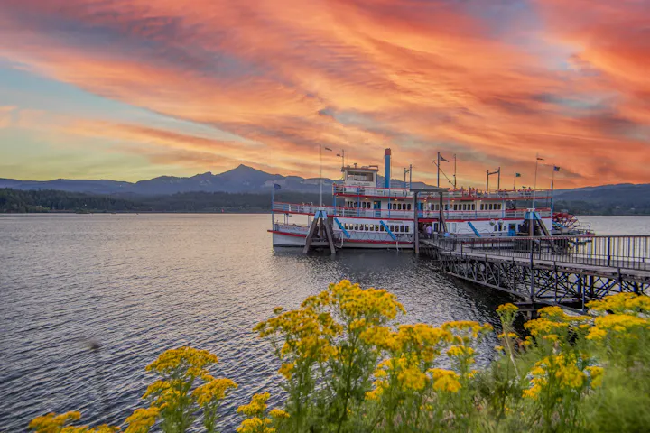 A colorful sunset over a lake with a vintage boat docked at a wooden pier, surrounded by flowers and mountains in the background.