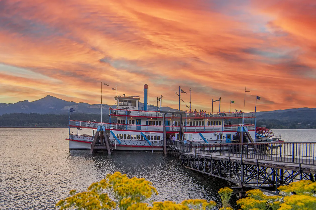 A colorful sunset over a lake with a vintage boat docked at a wooden pier, surrounded by flowers and mountains in the background.
