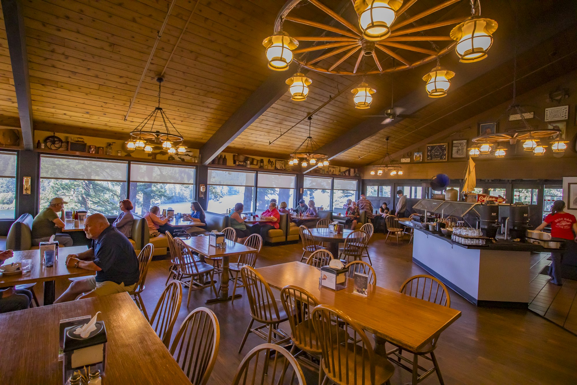 Cozy restaurant interior with wooden tables, diners seated by large windows, and wagon wheel chandeliers overhead.