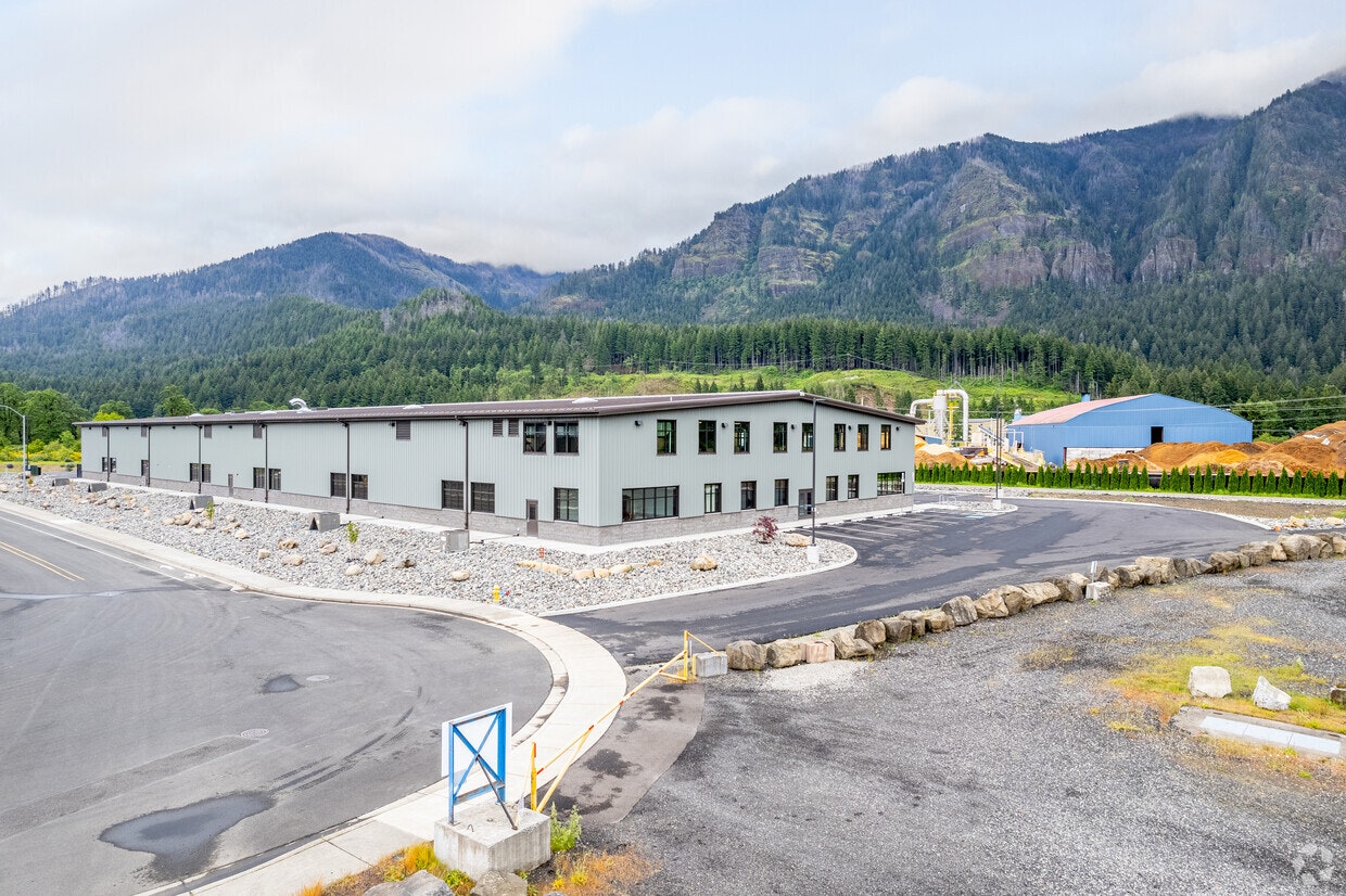 A modern industrial building near mountains, surrounded by gravel and landscaped areas, with a road curving around it.