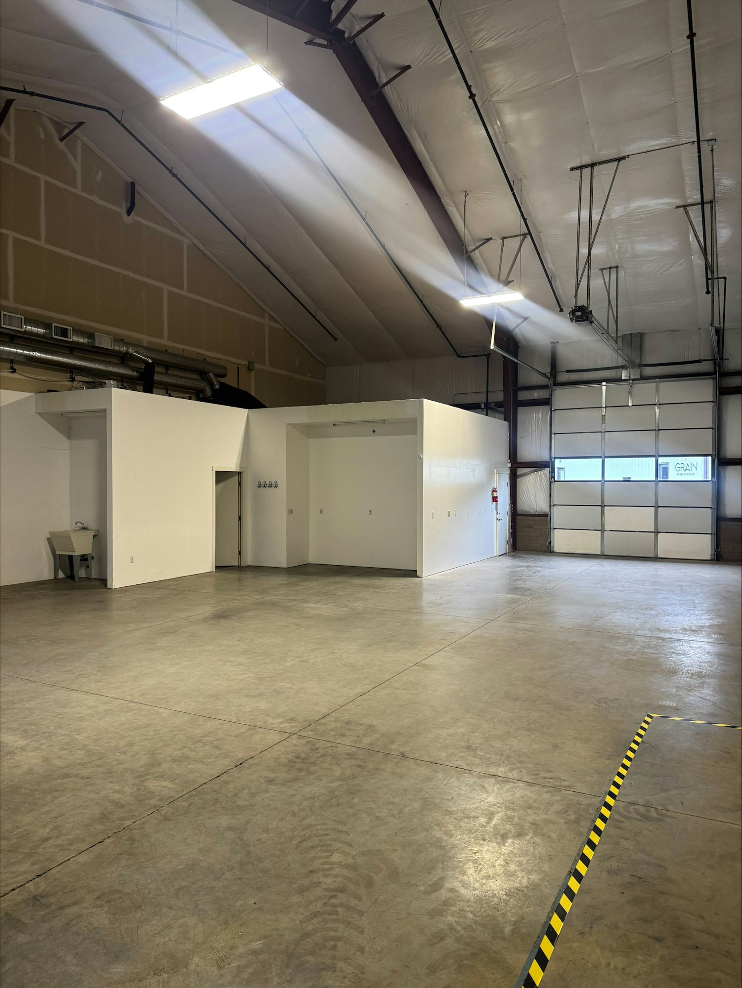 The image shows an empty industrial space with concrete floors, high ceilings, and some white walls, illuminated by overhead lights.