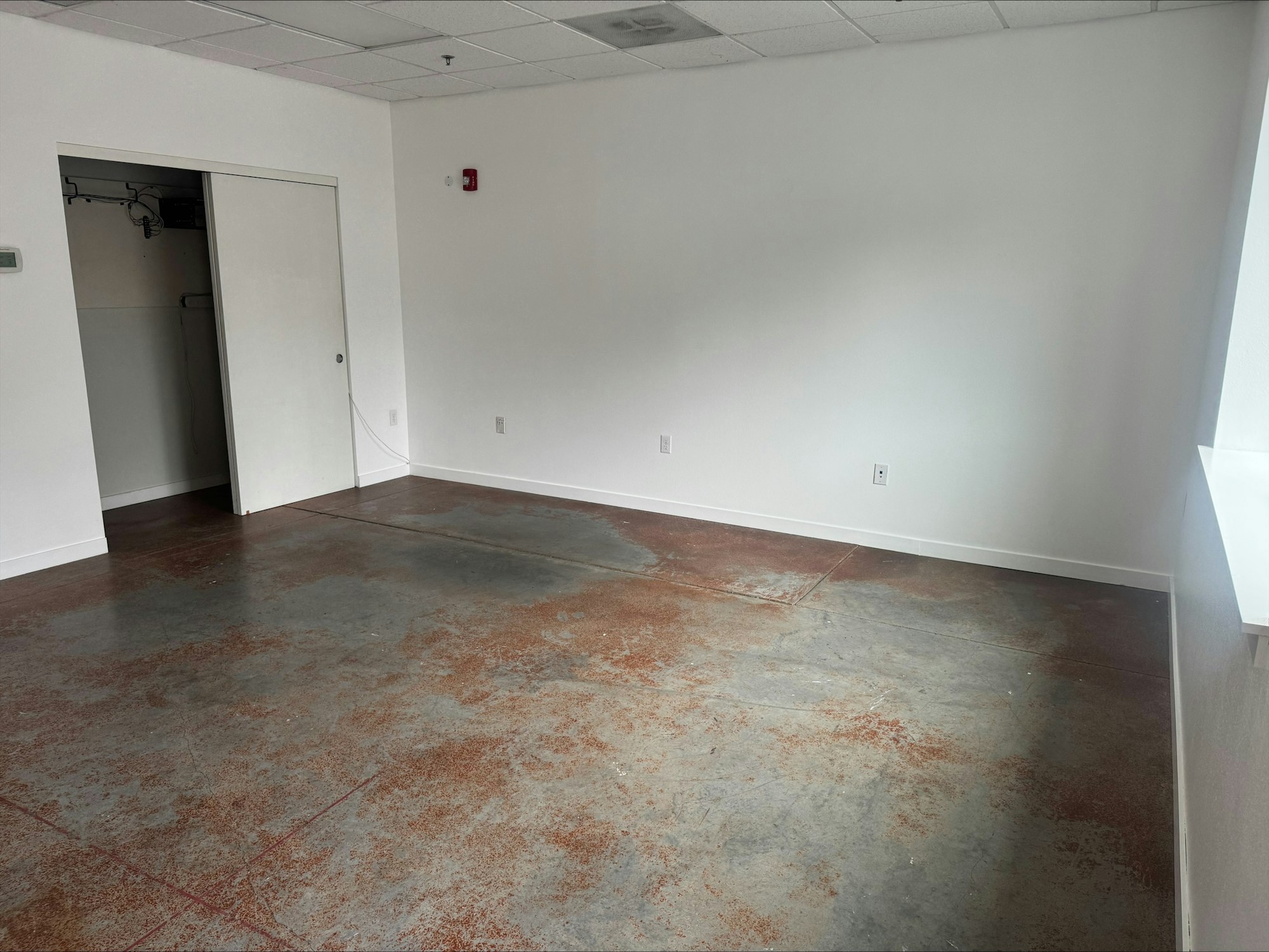 The image shows a sparsely furnished room with a polished concrete floor and white walls, including a closet and window.