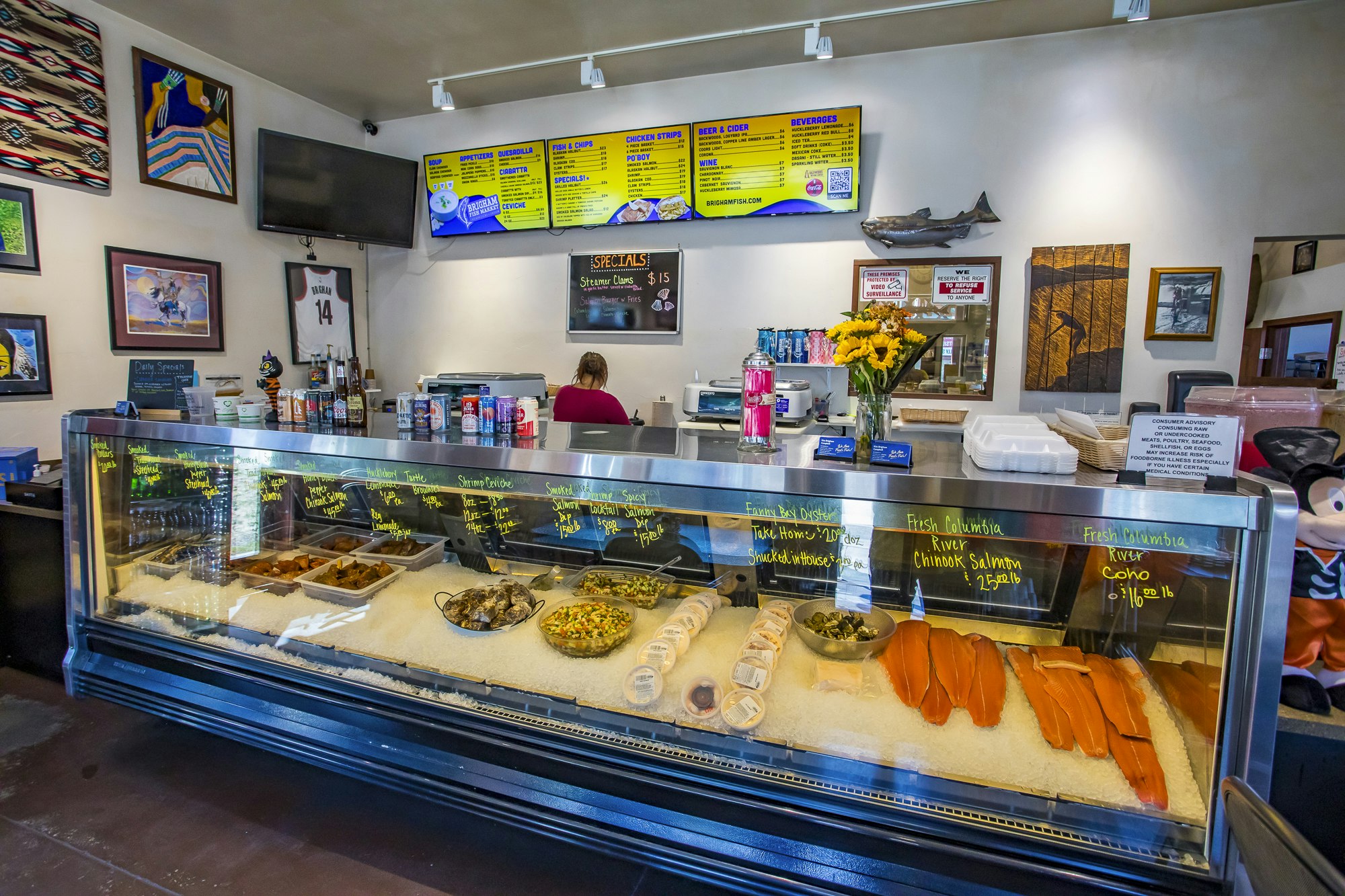 A seafood deli with various items displayed, menu boards above, flowers, and framed wall art.