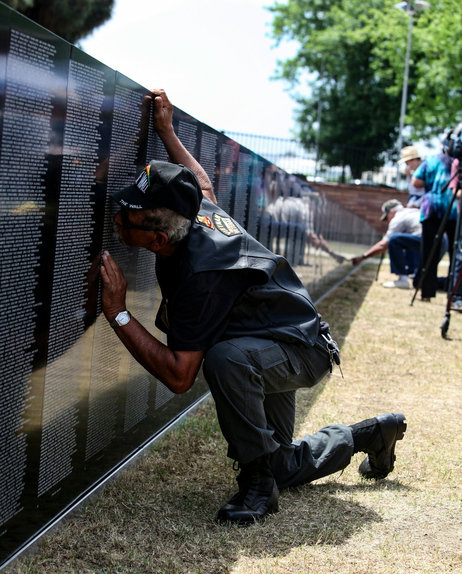 A veteran kneels by a memorial wall, touching the names, evoking reflection and remembrance. Others are present in the background.