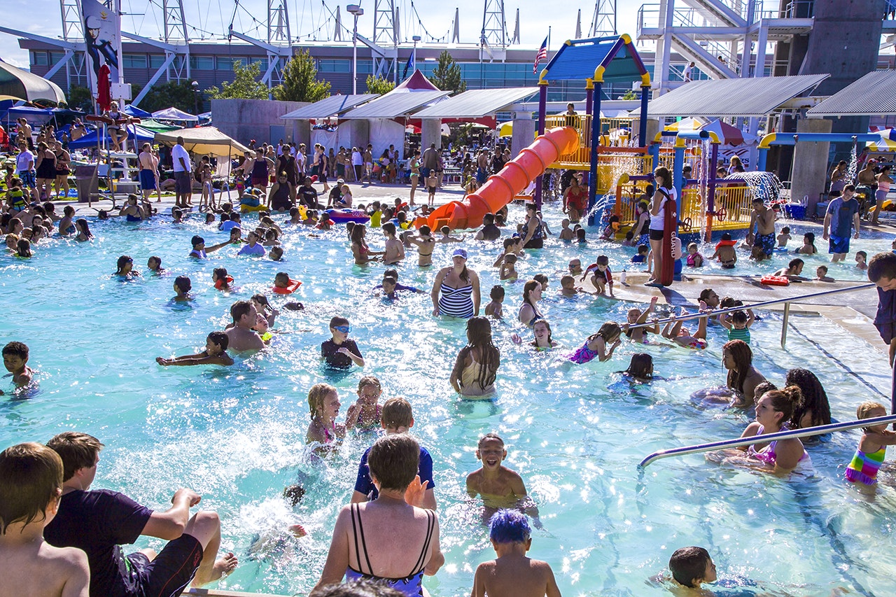A crowded outdoor swimming pool with people enjoying and a waterslide in the background.