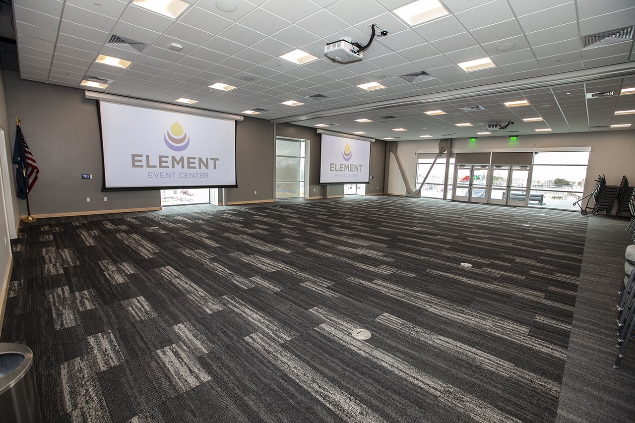 An empty event center room with patterned carpet, projectors, a presentation screen, American flag, and windows.